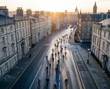 Vue aérienne d’Édimbourg au lever du jour, avec un groupe de cyclistes roulant sur une route urbaine dans une ambiance de grande course cycliste.