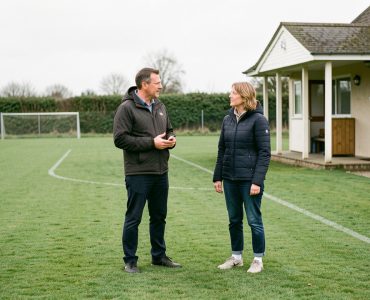 Terrain de football amateur vide avec un adulte référent échangeant calmement avec une parente près du club-house, dans une ambiance protectrice et sereine.